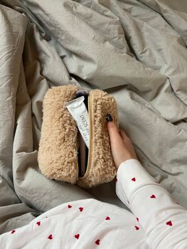 A cozy scene of a woman's hand holding a fluffy makeup bag on bed sheets.