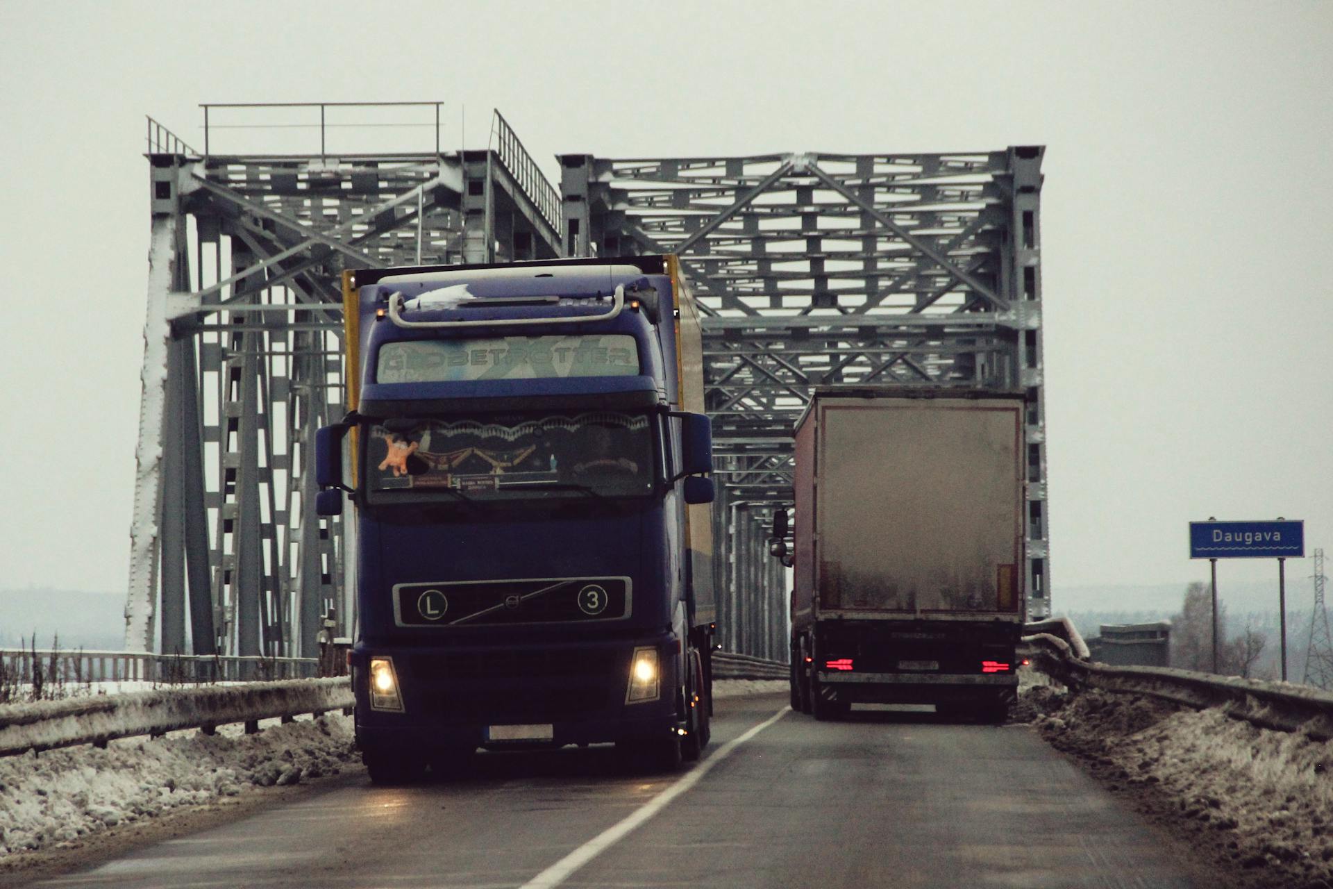 Two trucks navigate a snowy bridge over the Daugava River in Latvia during winter.
