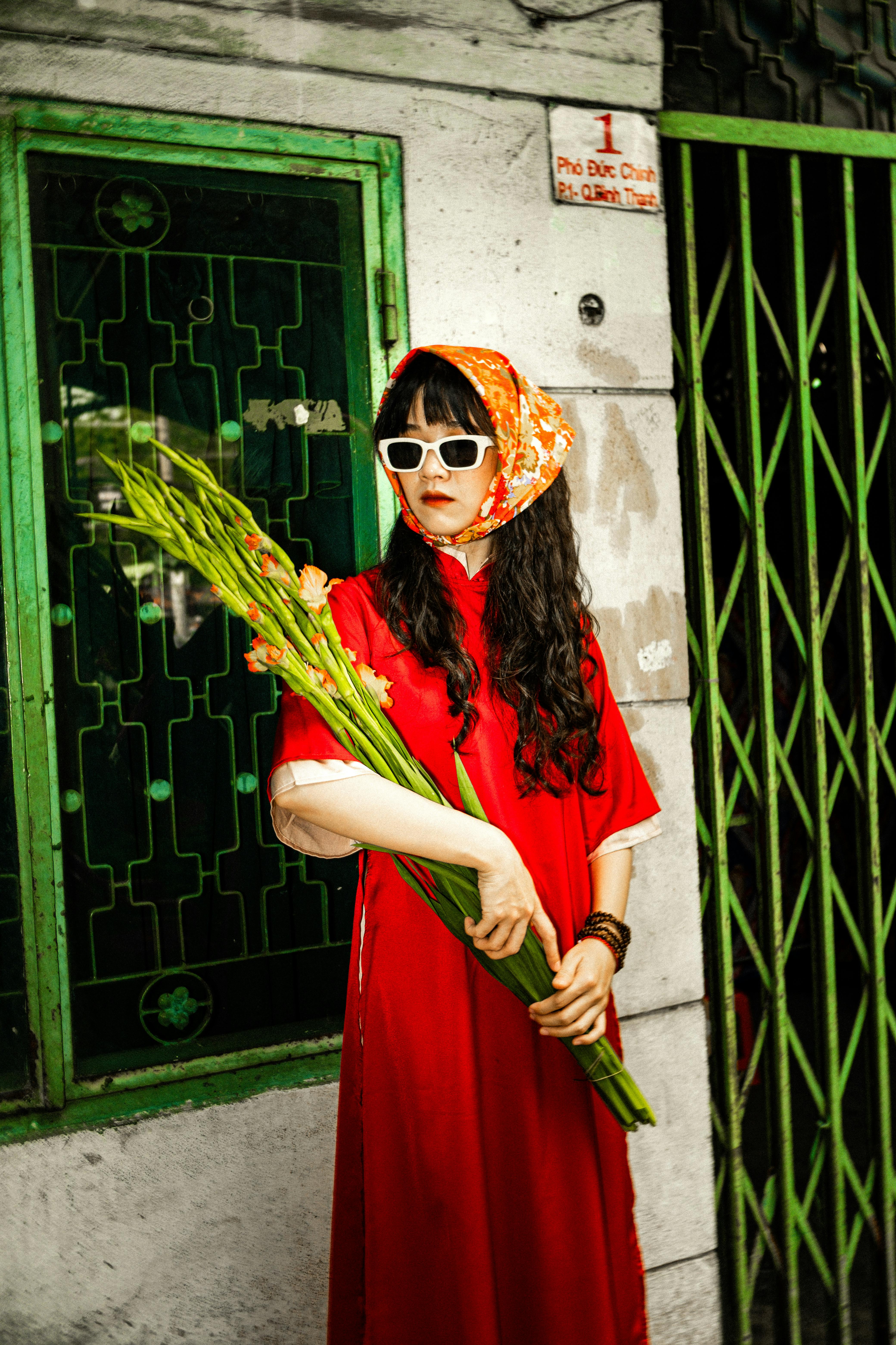 Fashionable woman in a red dress and sunglasses holding flowers in an urban setting with green accents.