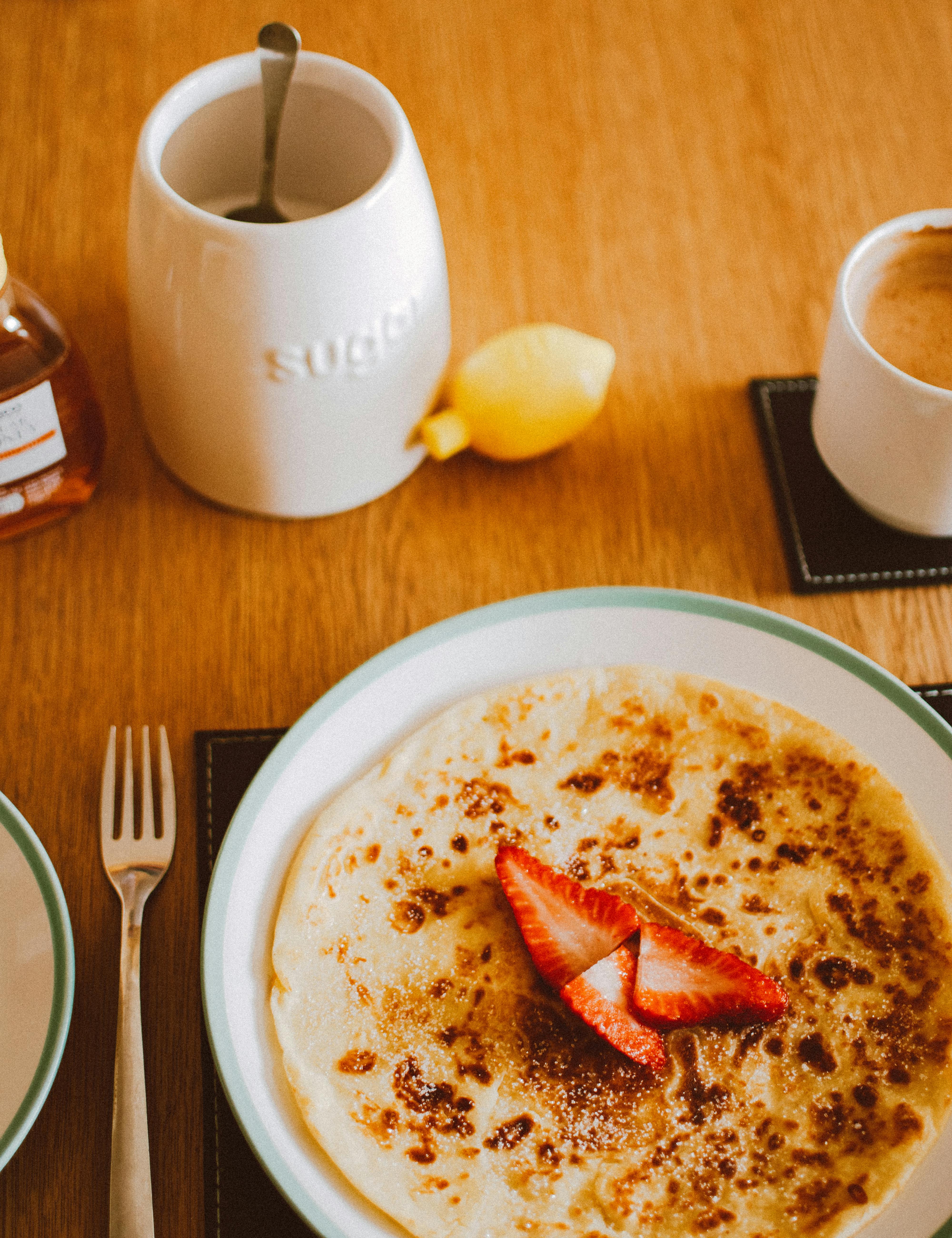 Close-up of breakfast crepes topped with fresh strawberries, accompanied by coffee and sugar jar.