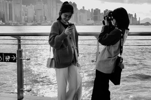 Women Standing by Railing on Sea Shore and Taking Pictures in Black and White