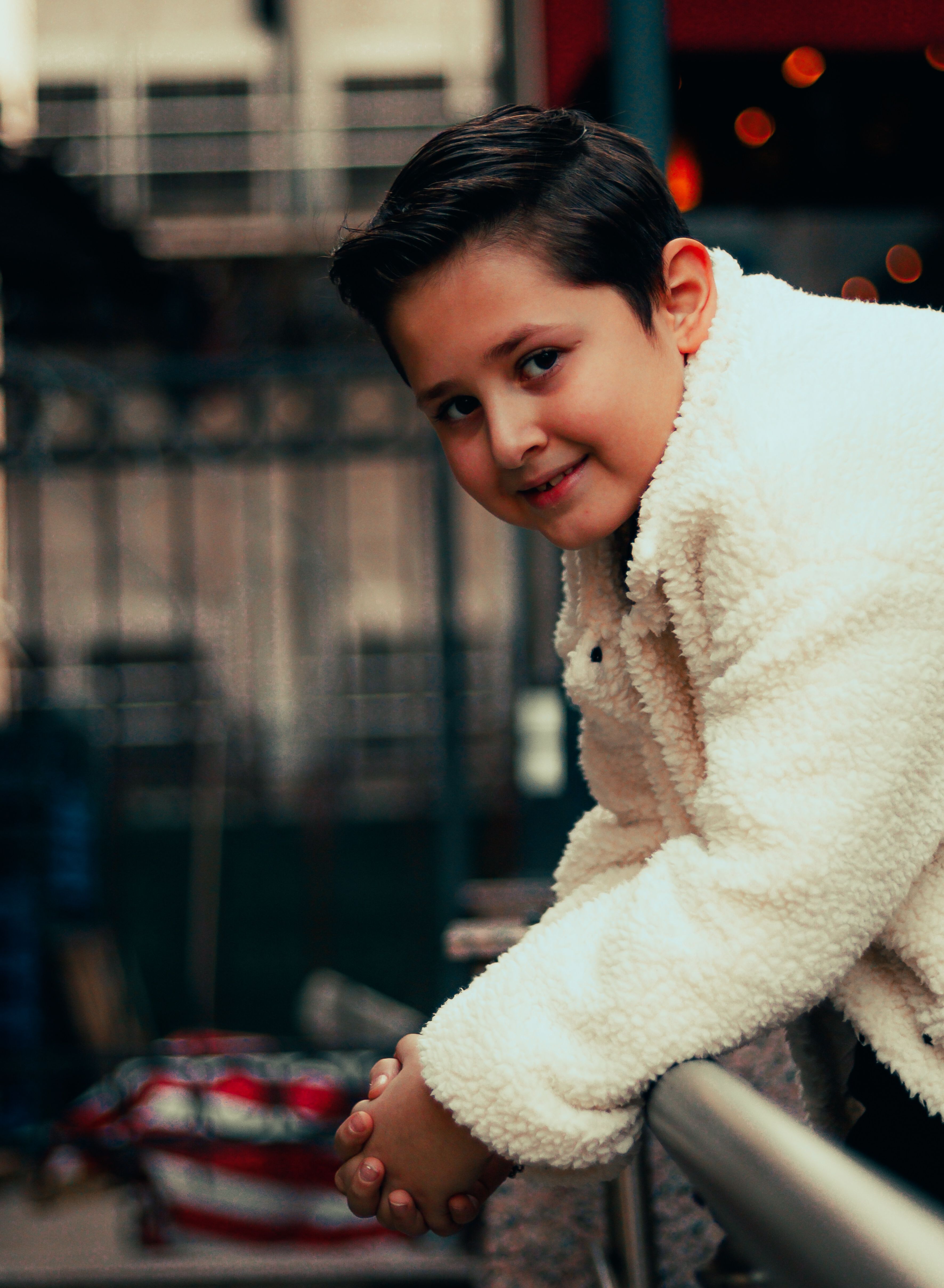 Portrait of a young boy in a white jacket leaning on a railing, with a city backdrop.