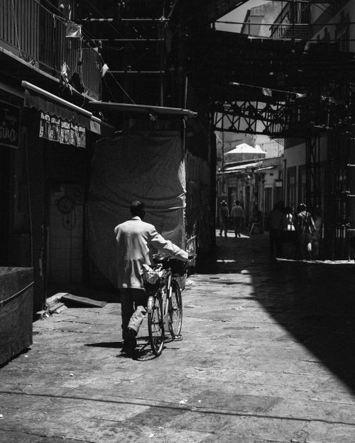 Free Black and white photo of a man walking with a bicycle in a narrow urban alley during the day. Stock Photo