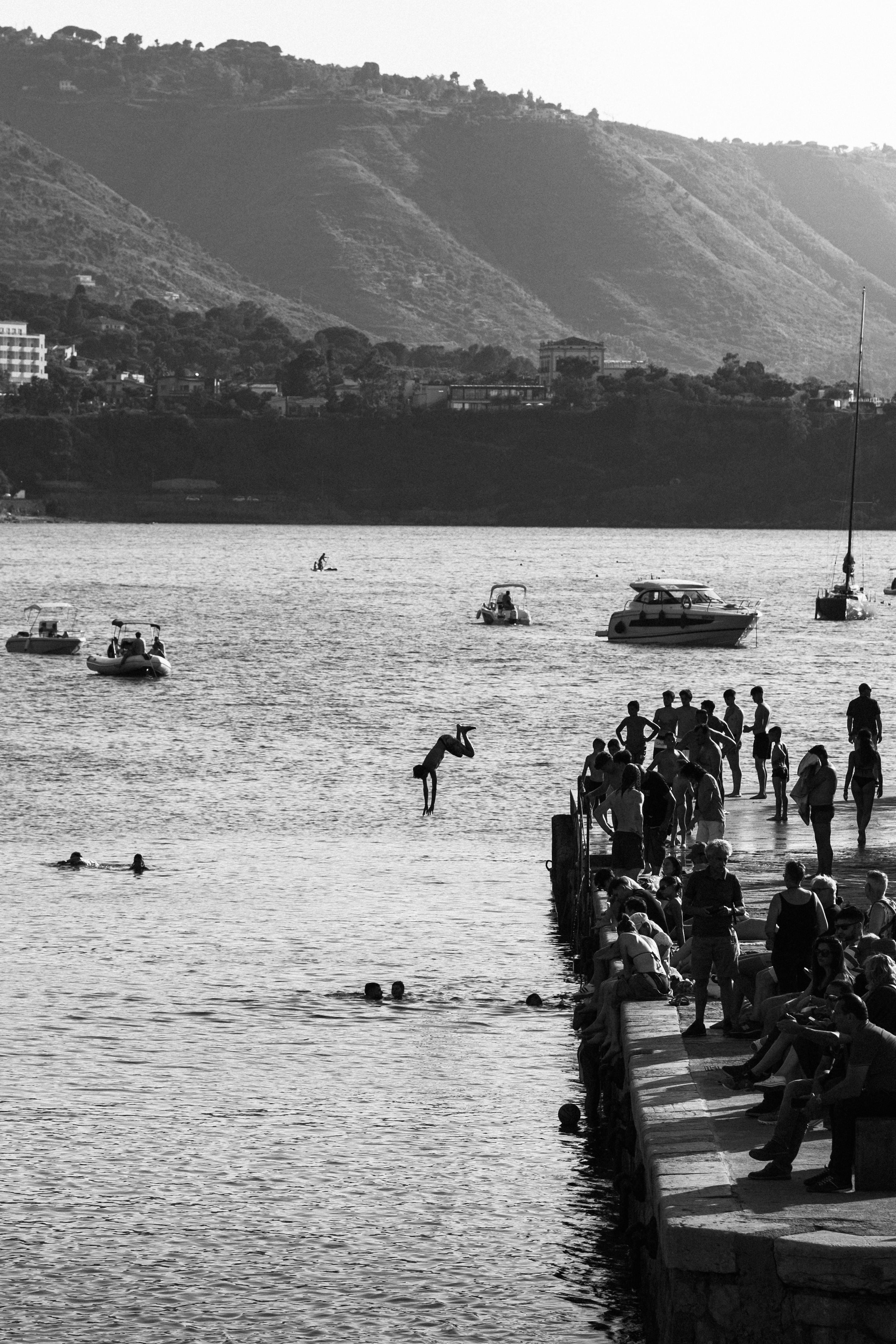 Black and white photo of a summer pier with people diving and boats in the background.
