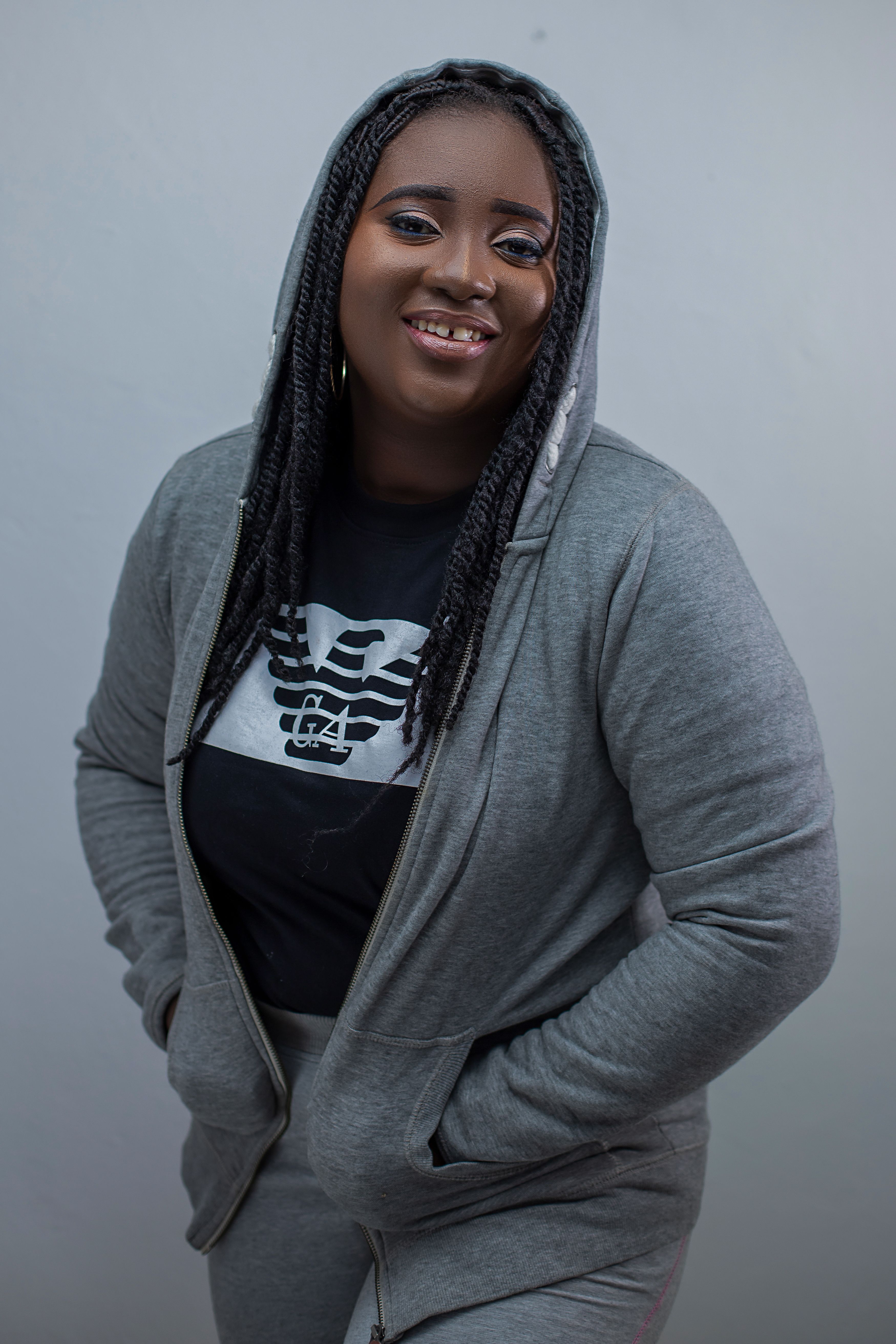 Fashion portrait of a smiling woman in a gray hoodie with braided hair against a neutral background.