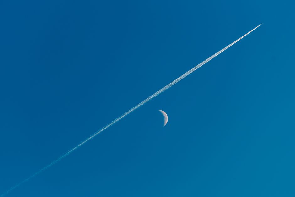 Dramatic capture of a contrail crossing a crescent moon against a clear blue sky.