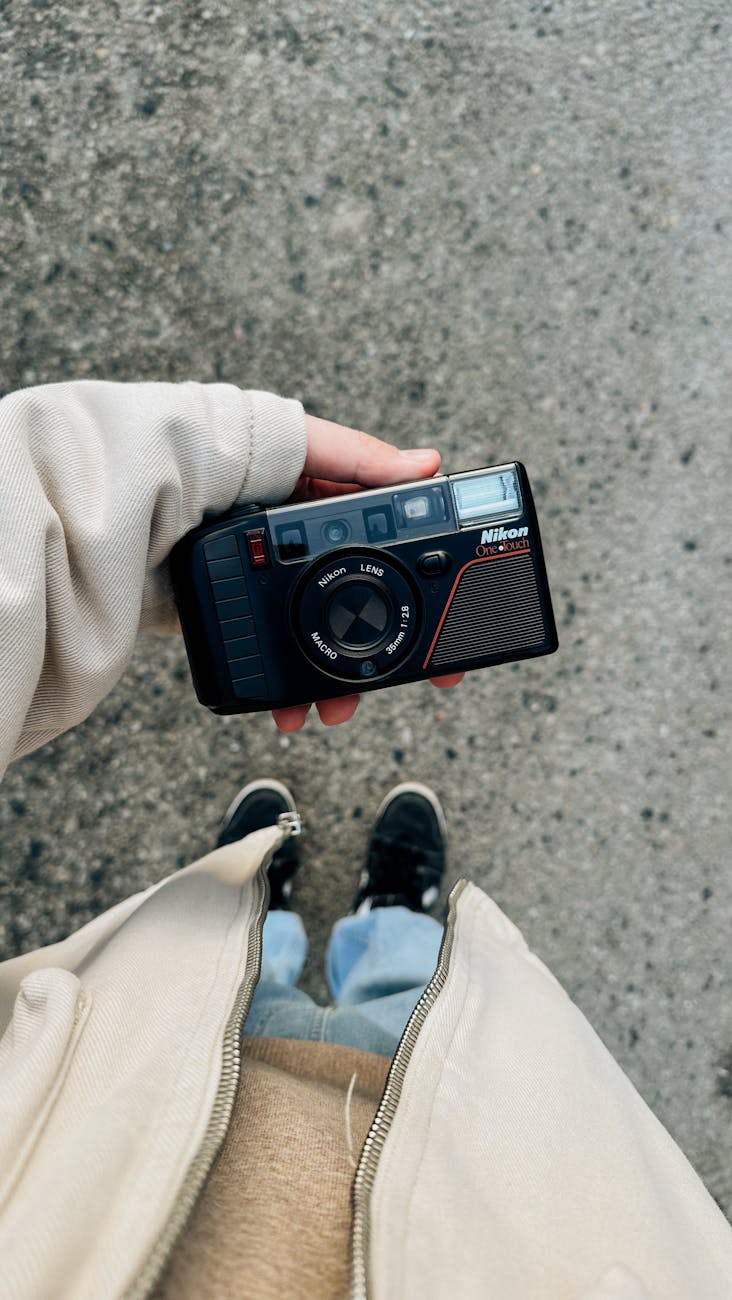 A top view shot of a person holding a vintage camera outdoors, wearing a beige jacket and sneakers.