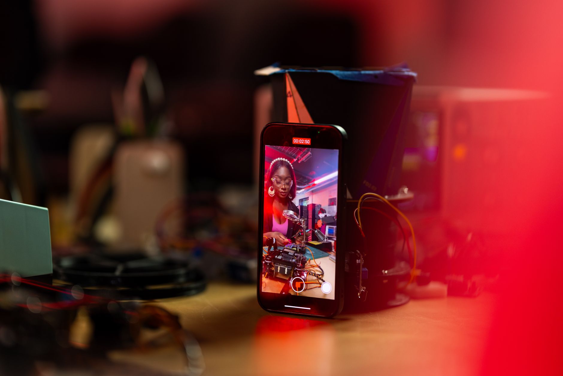 A woman records a tech tutorial using her smartphone on a desk filled with electronics.