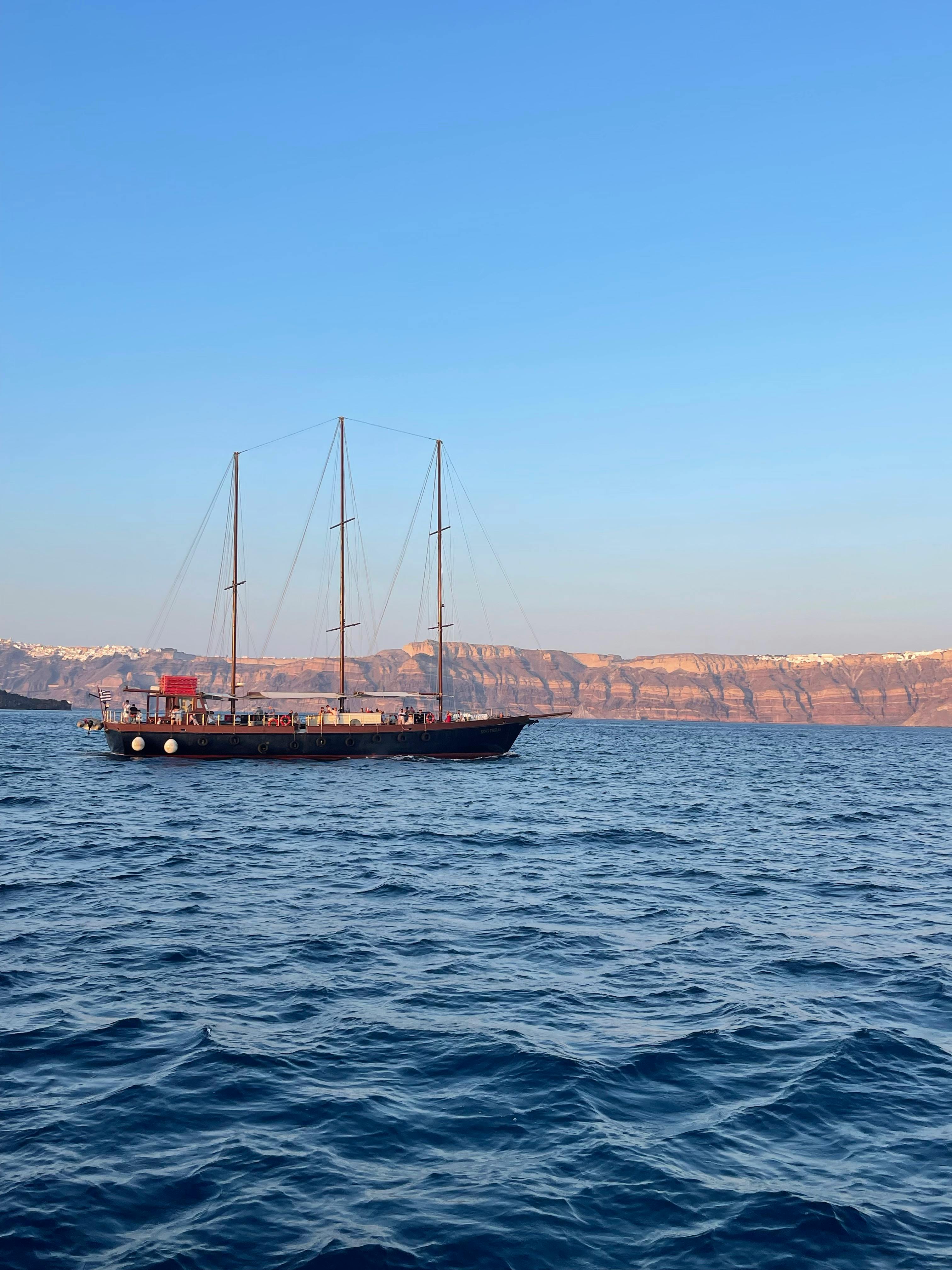 A sailing ship cruises on the Aegean Sea with Santorini cliffs in the background.
