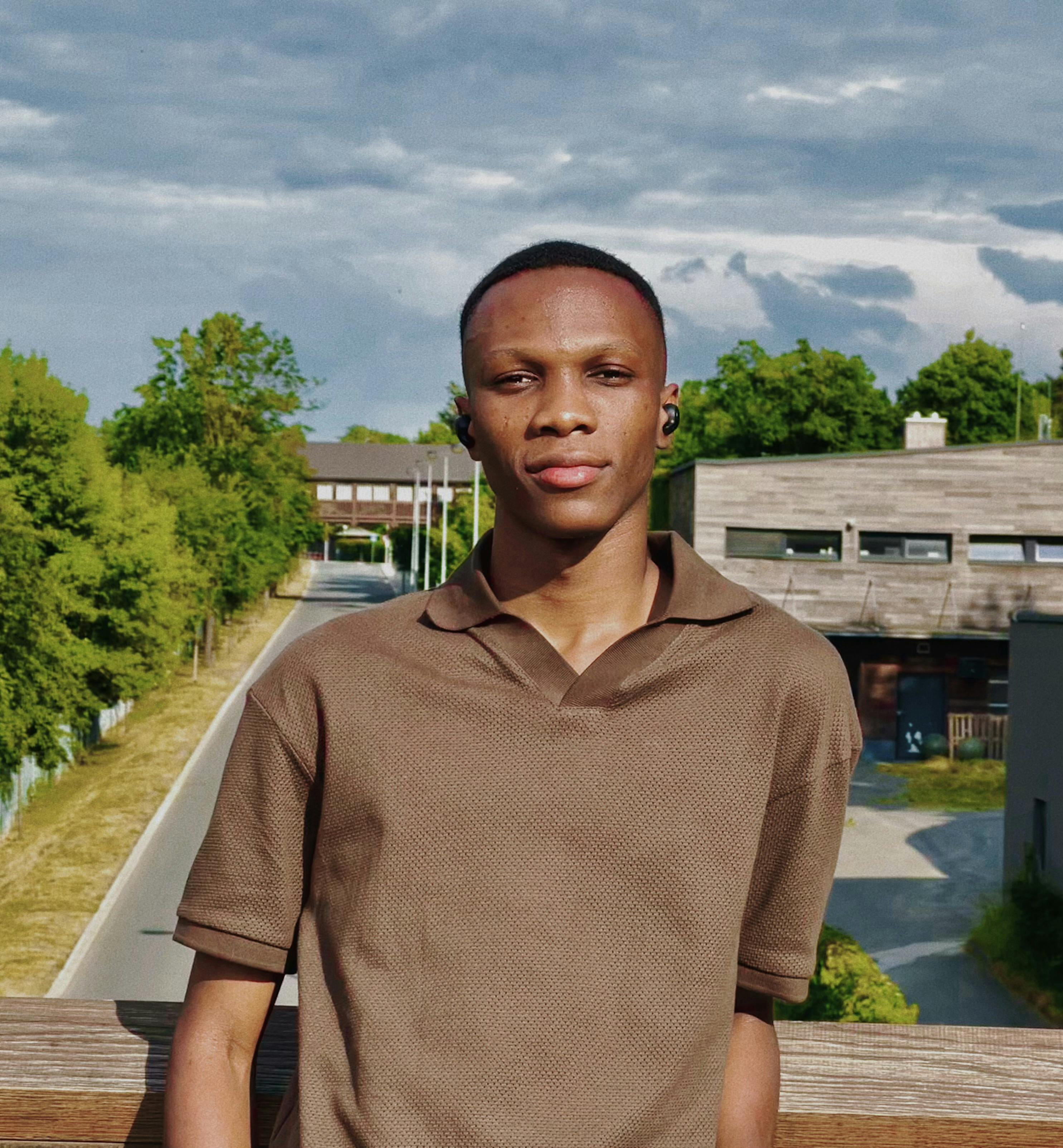 Portrait of a man standing on a bridge with lush greenery and a cloudy sky.