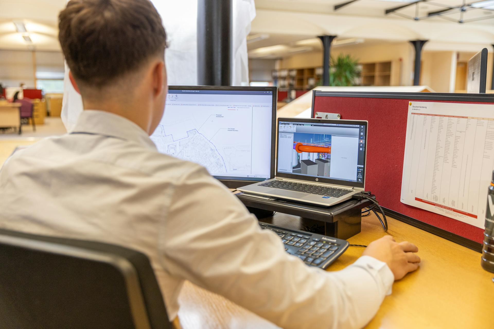 Back view of engineer focused on CAD design at dual monitors in bright office setting.