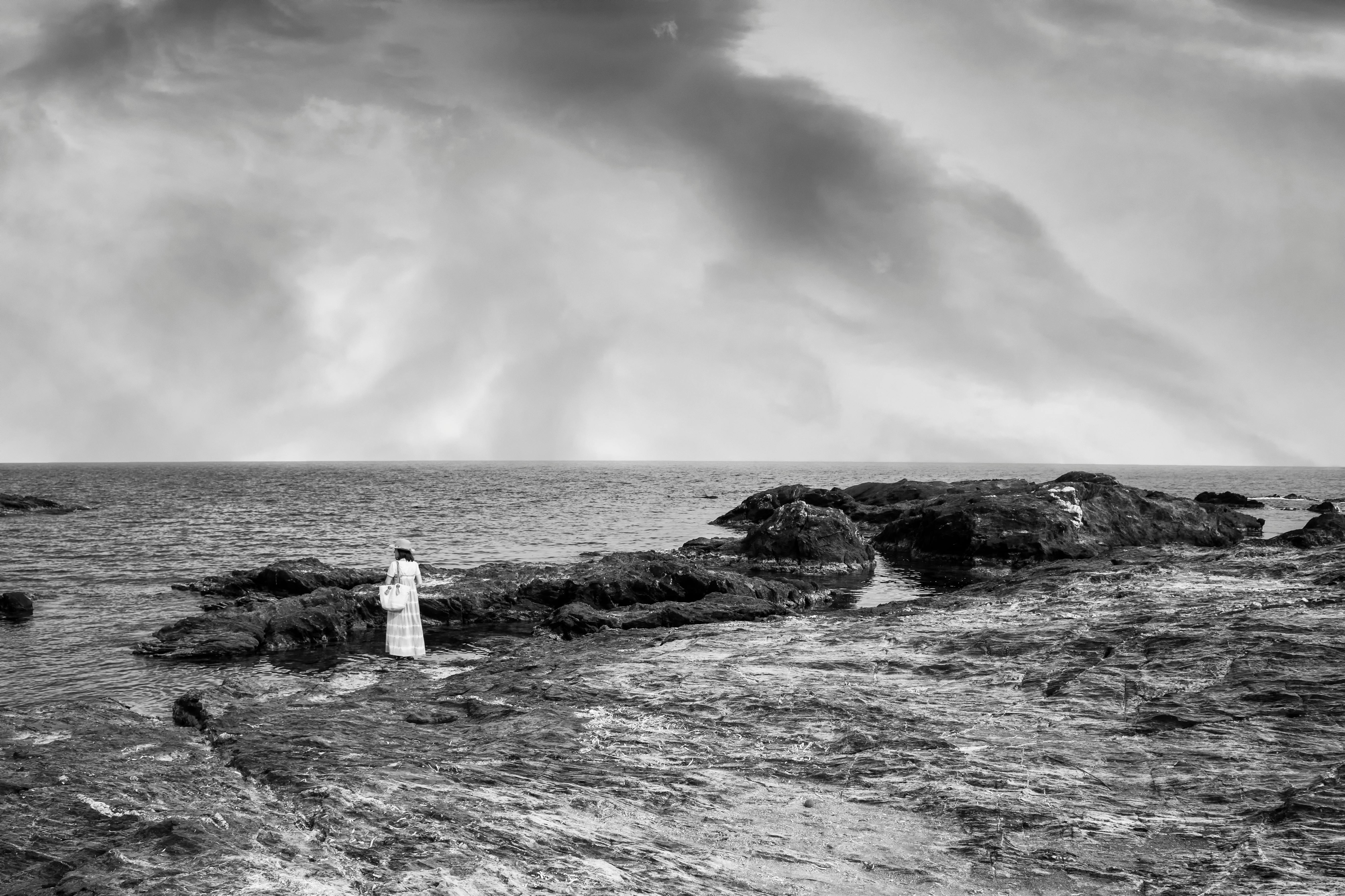 A serene black and white seascape featuring a woman standing on rocky terrain by the ocean.