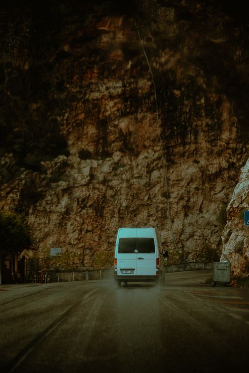 Free A white mini van travels on a winding road beside a steep rocky cliff, capturing the essence of travel and adventure. Stock Photo