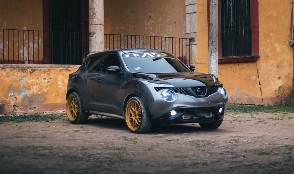A sleek gray Nissan Juke with golden rims parked near a rustic building exterior.