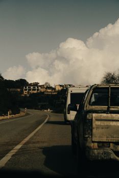 Cars traveling on a rural road, heading towards a small village under a cloudy sky.
