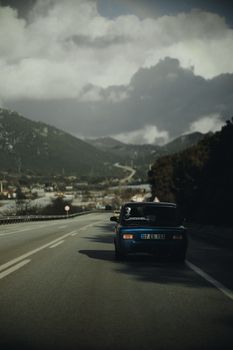 A vintage car travels on a scenic mountain road under dramatic clouds.