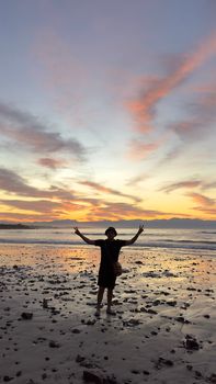 Silhouette of a man with arms raised, standing on a rocky beach during sunset.