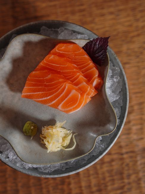 Glossy orange-pink salmon sashimi sliced into thick diagonal pieces arranged on a black plate with pickled ginger and a small dish of soy sauce