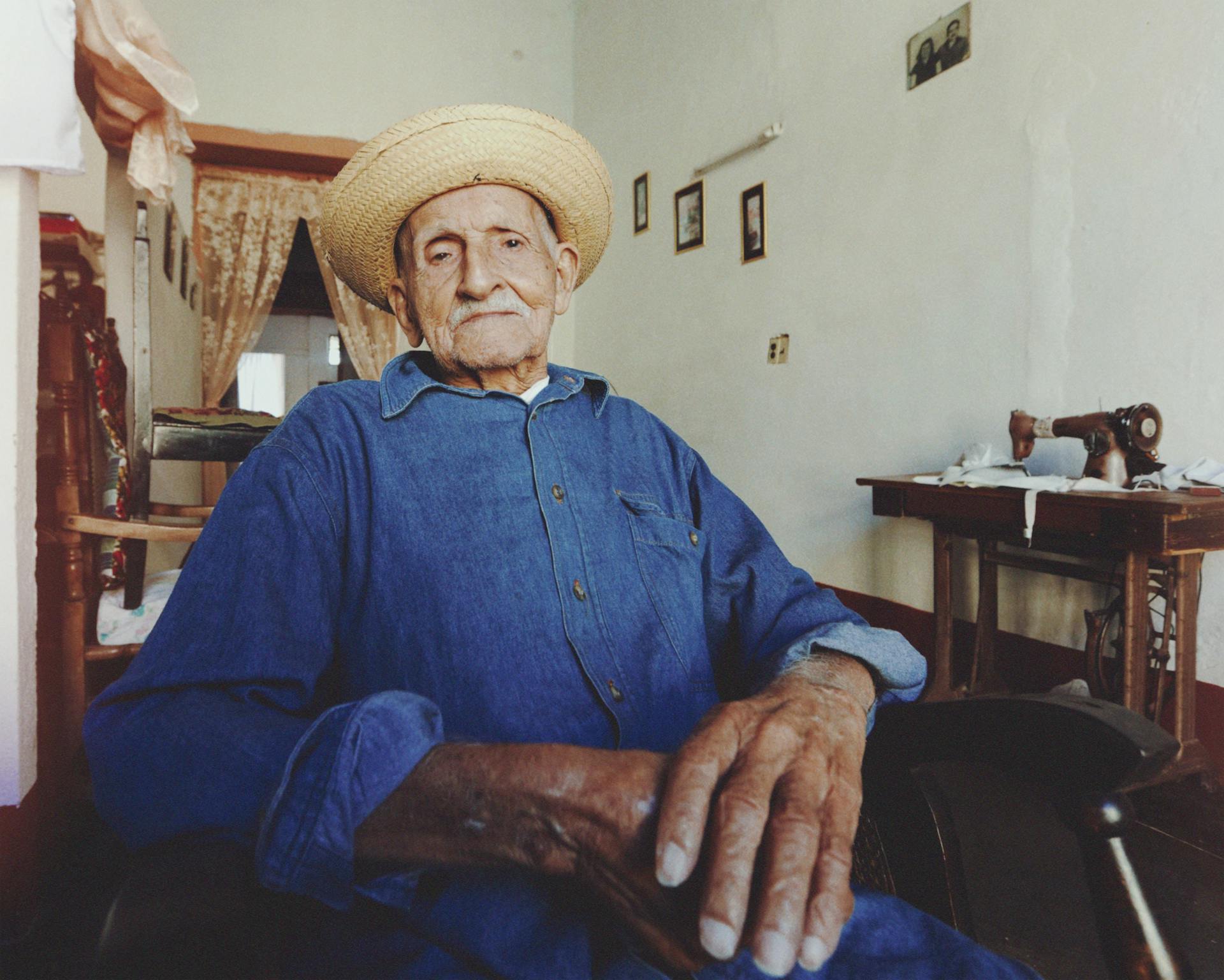 Elderly man in denim and straw hat sitting indoors, portraying the essence of retirement and vintage lifestyle.