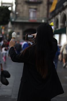 Woman with long hair holding ice cream, capturing urban scene with smartphone.