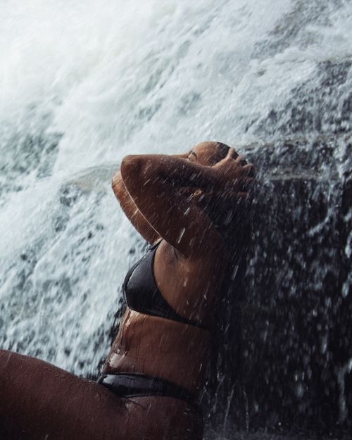 Free A woman in a bikini sitting under a waterfall, expressing a serene moment in nature. Stock Photo