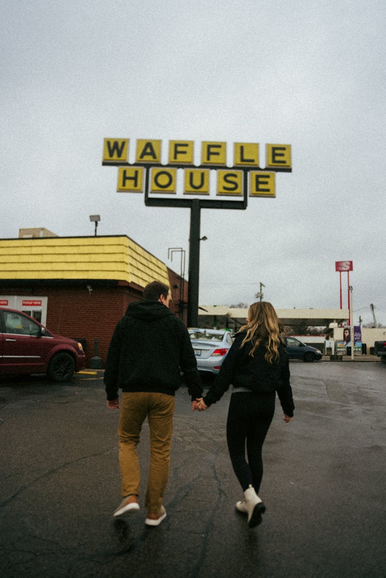 Man And Woman Holding Hands While Walking Through A Parking Lot Toward A Restaurant 