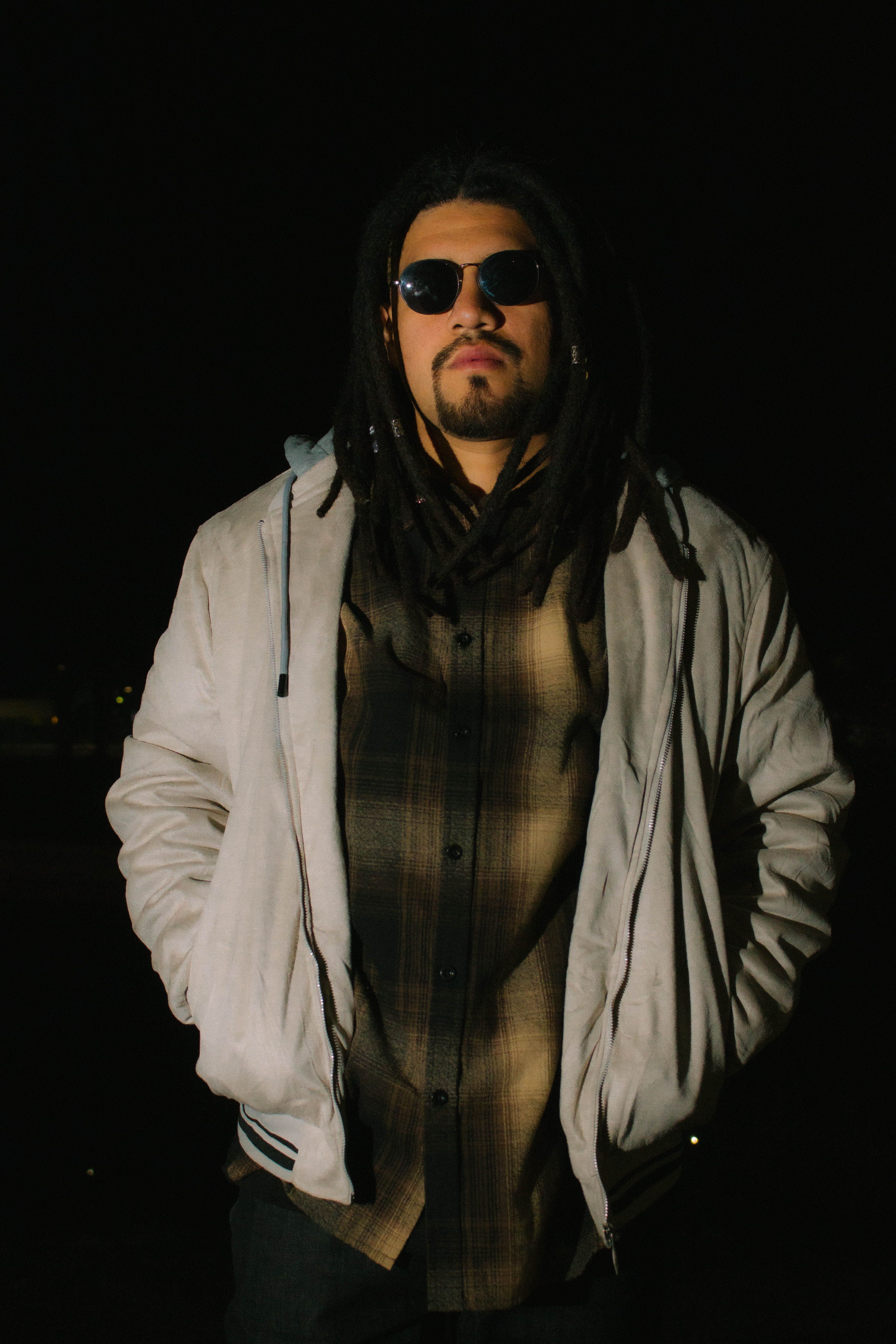 Portrait of a trendy man with dreadlocks wearing sunglasses and a jacket at night.