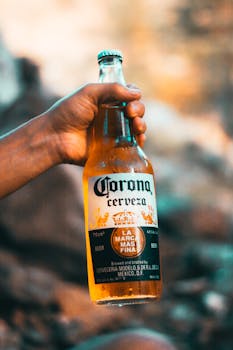 Close-up of a hand holding a Corona beer bottle outdoors, showcasing the brand label.