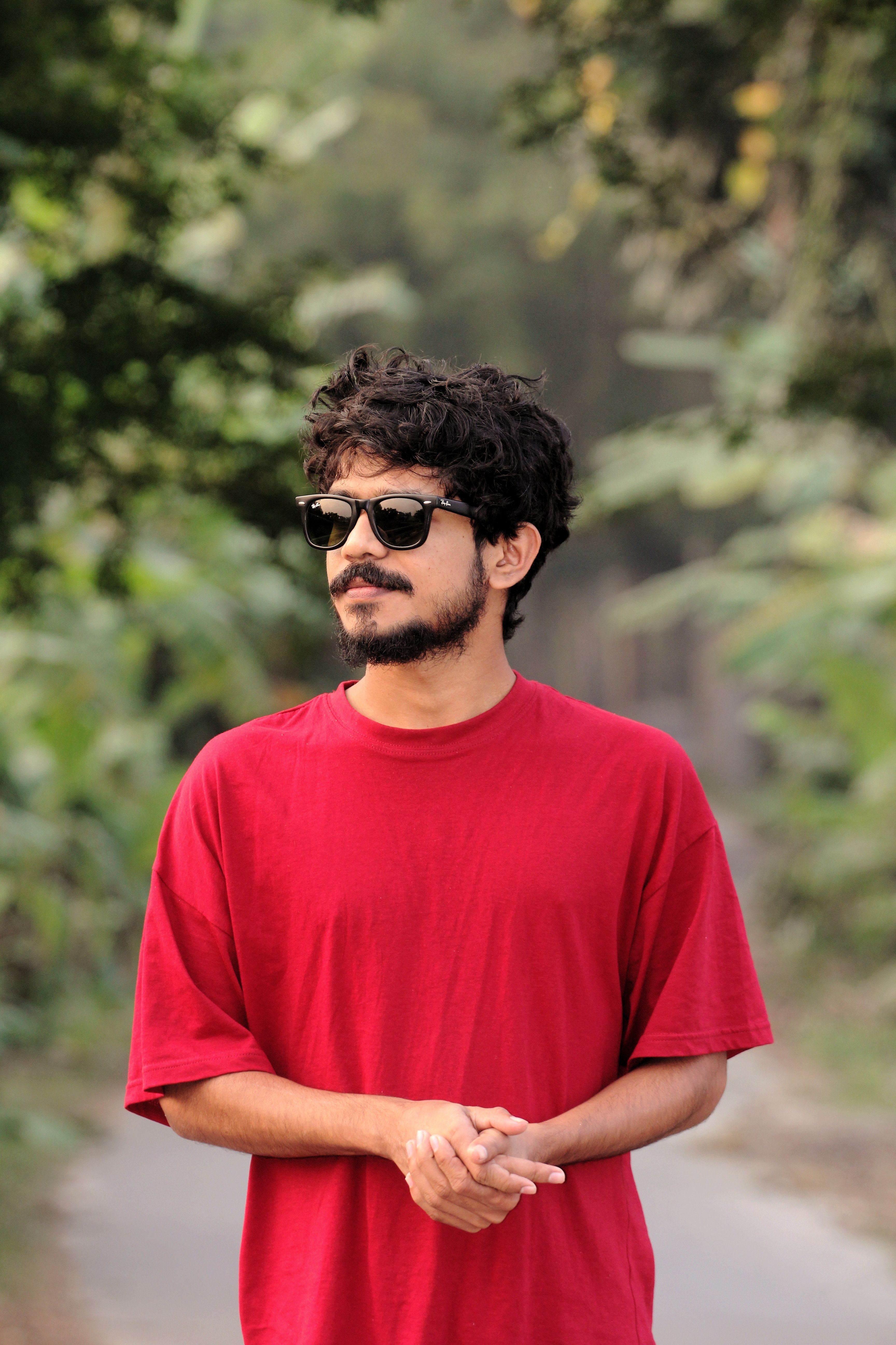 Portrait of a young man wearing sunglasses and a red t-shirt, standing outdoors in a lush green park setting.