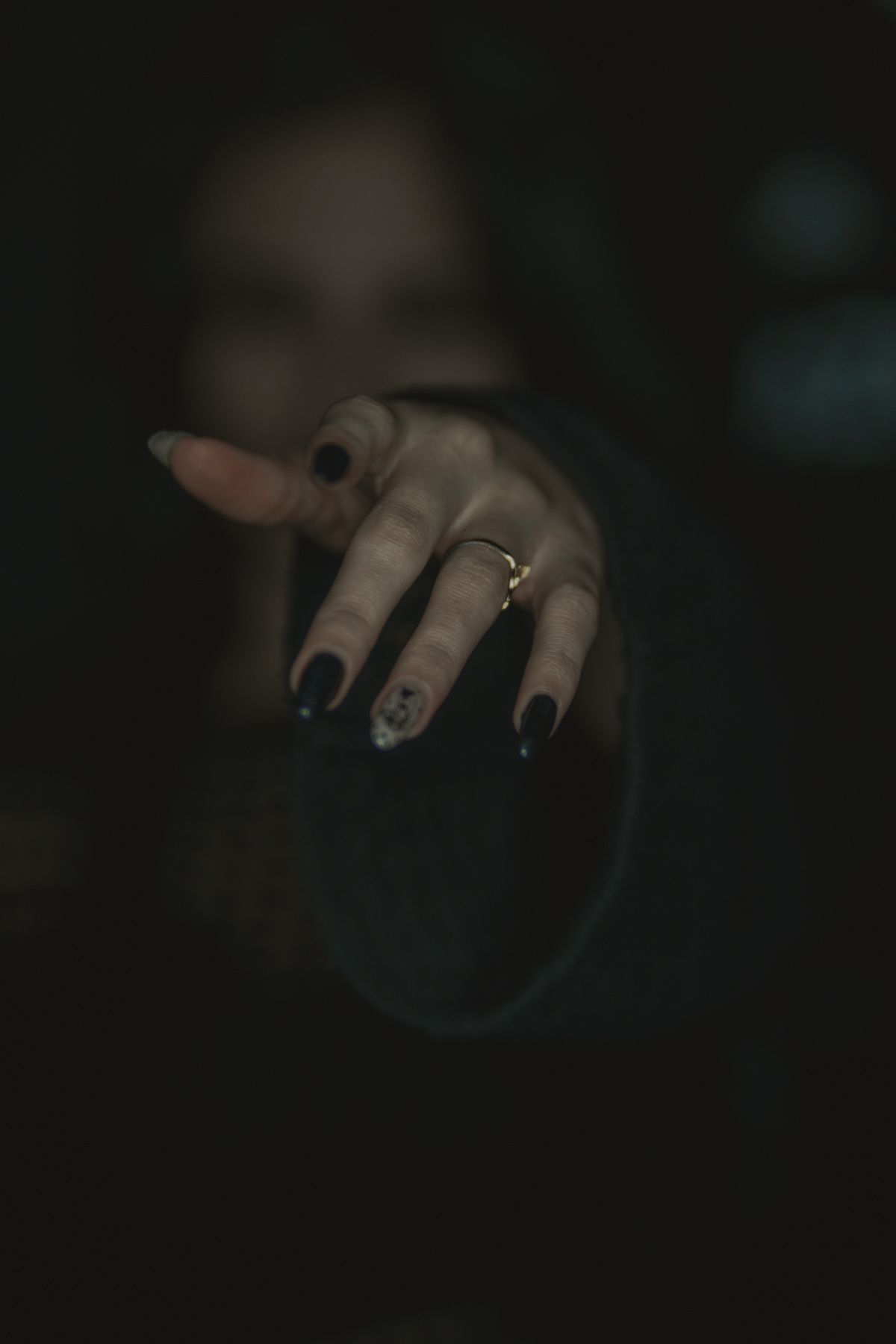 A dramatic focus on a woman's hand with dark nail polish and a ring, reaching forward in low light.