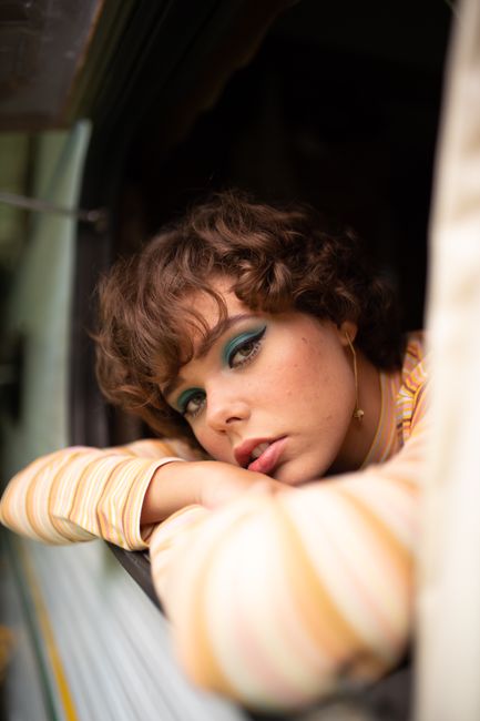 Portrait of a young woman with green eyeshadow and curly hair leaning out of a campervan window.