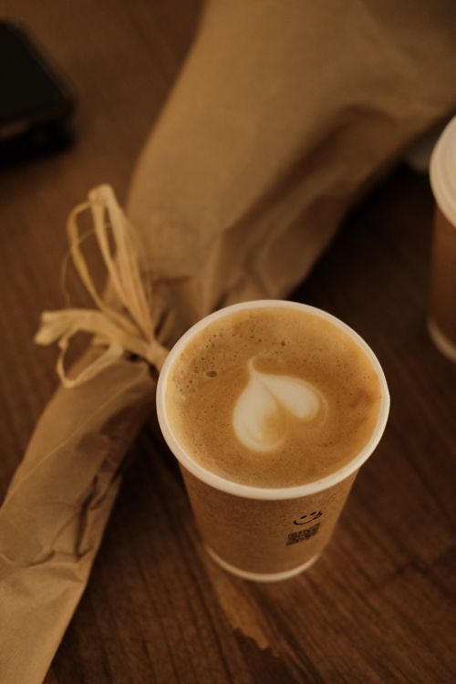 Free Warm coffee cup with heart latte art on a wooden table next to a wrapped bouquet. Stock Photo