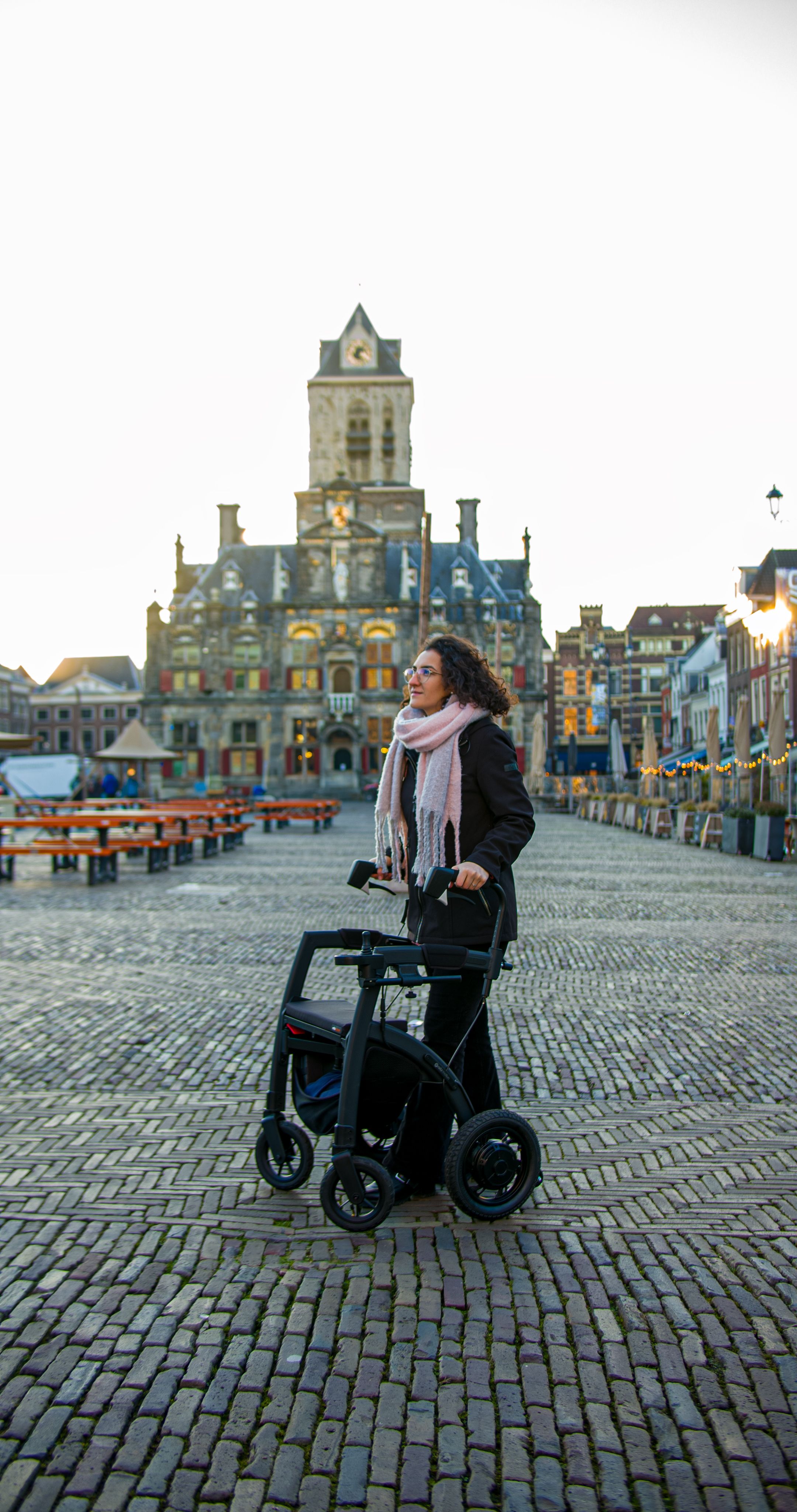 A woman with a walker strolls in the cobblestone square of Delft, Netherlands, in front of historic architecture.