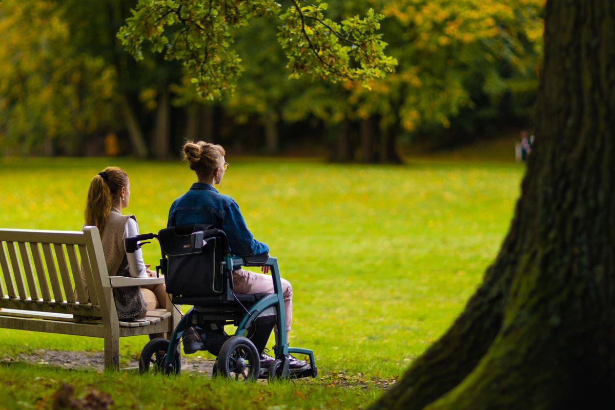 Two women sitting together in a park, one using a mobility chair