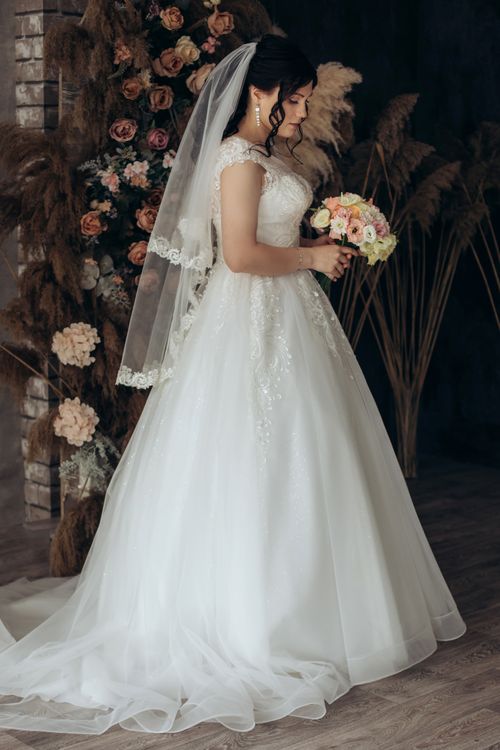 Free A beautiful bride elegantly holding a bouquet of flowers, posing in a classic wedding dress indoors. Stock Photo