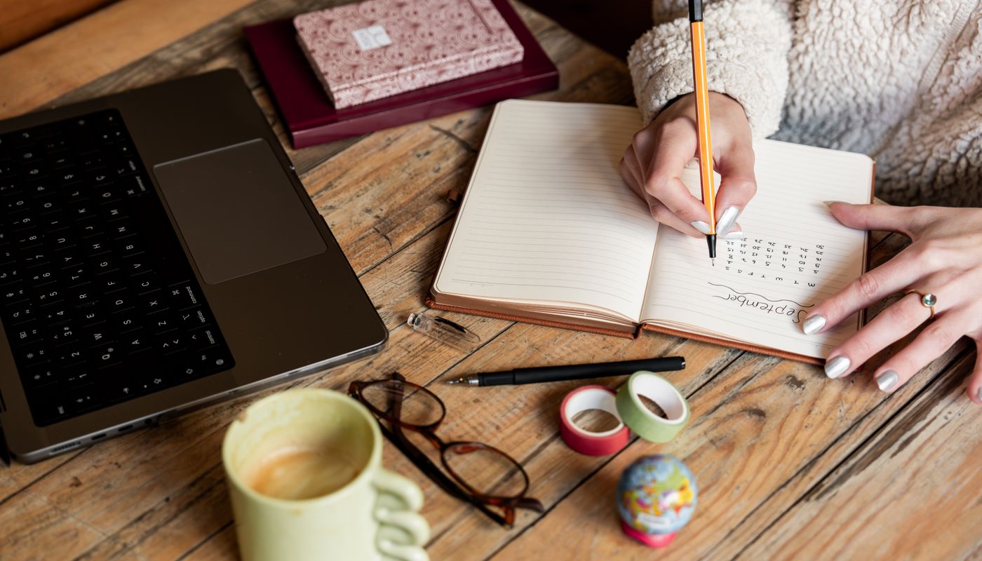 A desk with an open planner, pencil, coffee cup and laptop in warm light