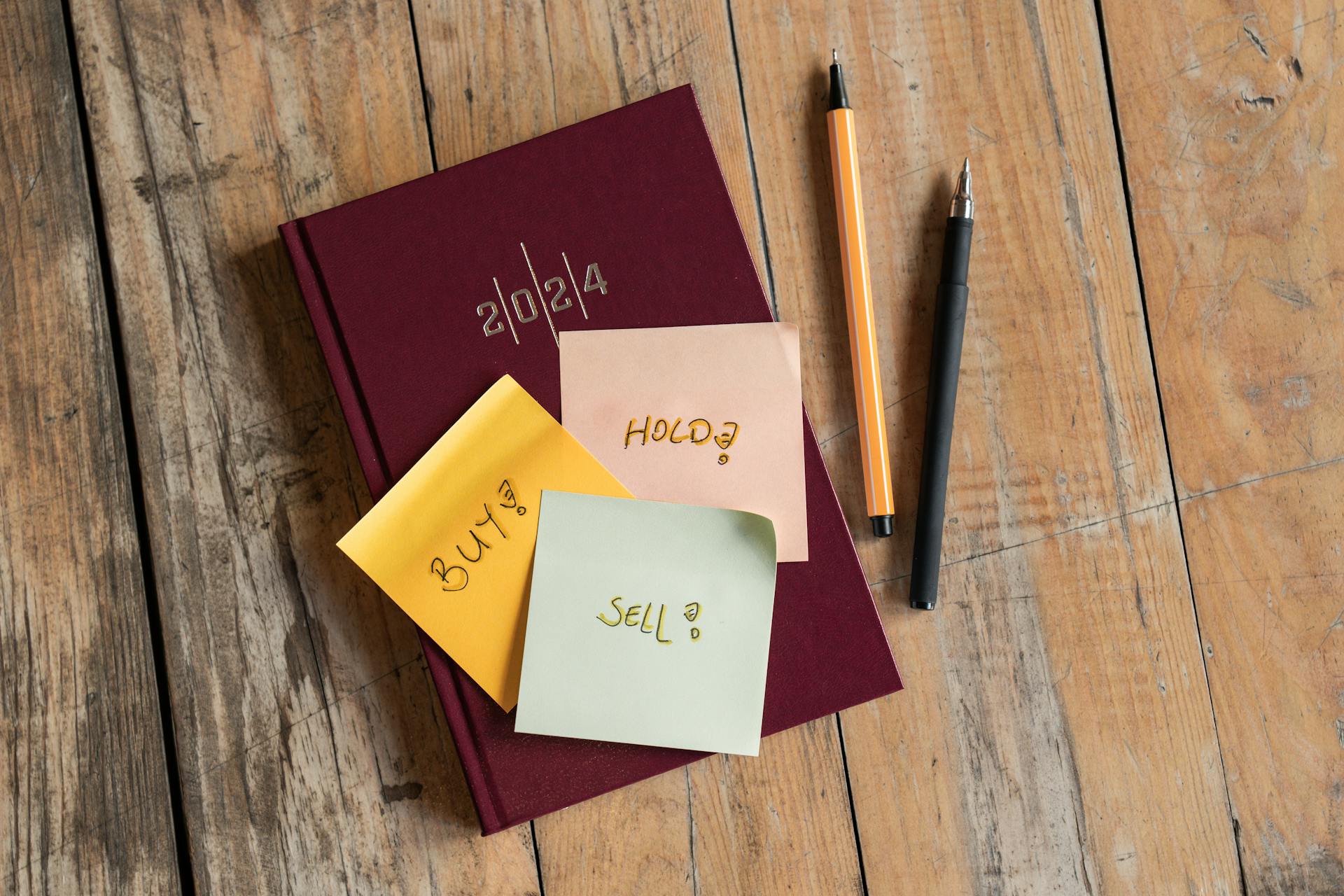 2024 planner with 'buy', 'hold', 'sell' notes and pens on a rustic wooden surface.