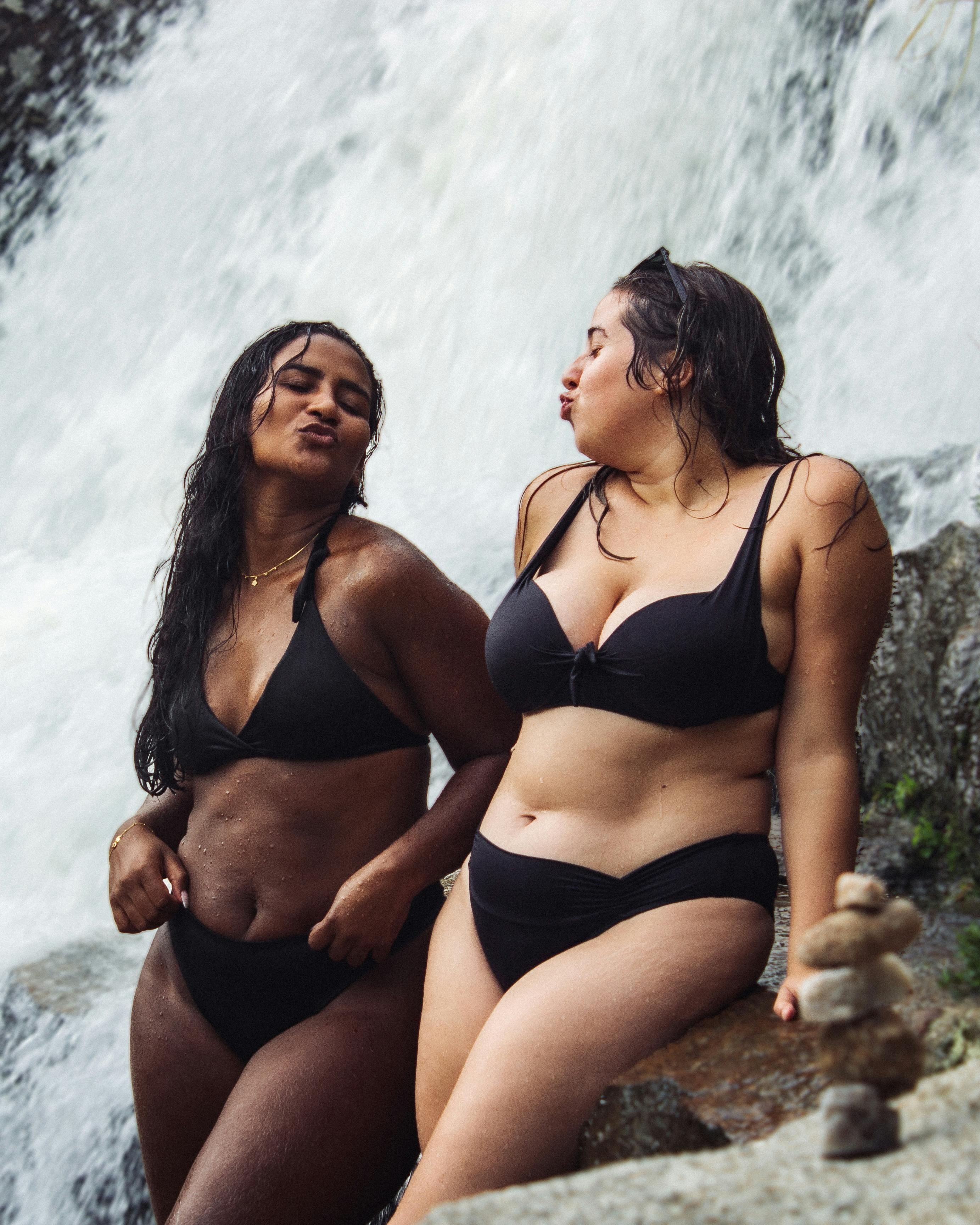 Free Two women in black bikinis posing playfully by a waterfall, exuding relaxation and joy. Stock Photo