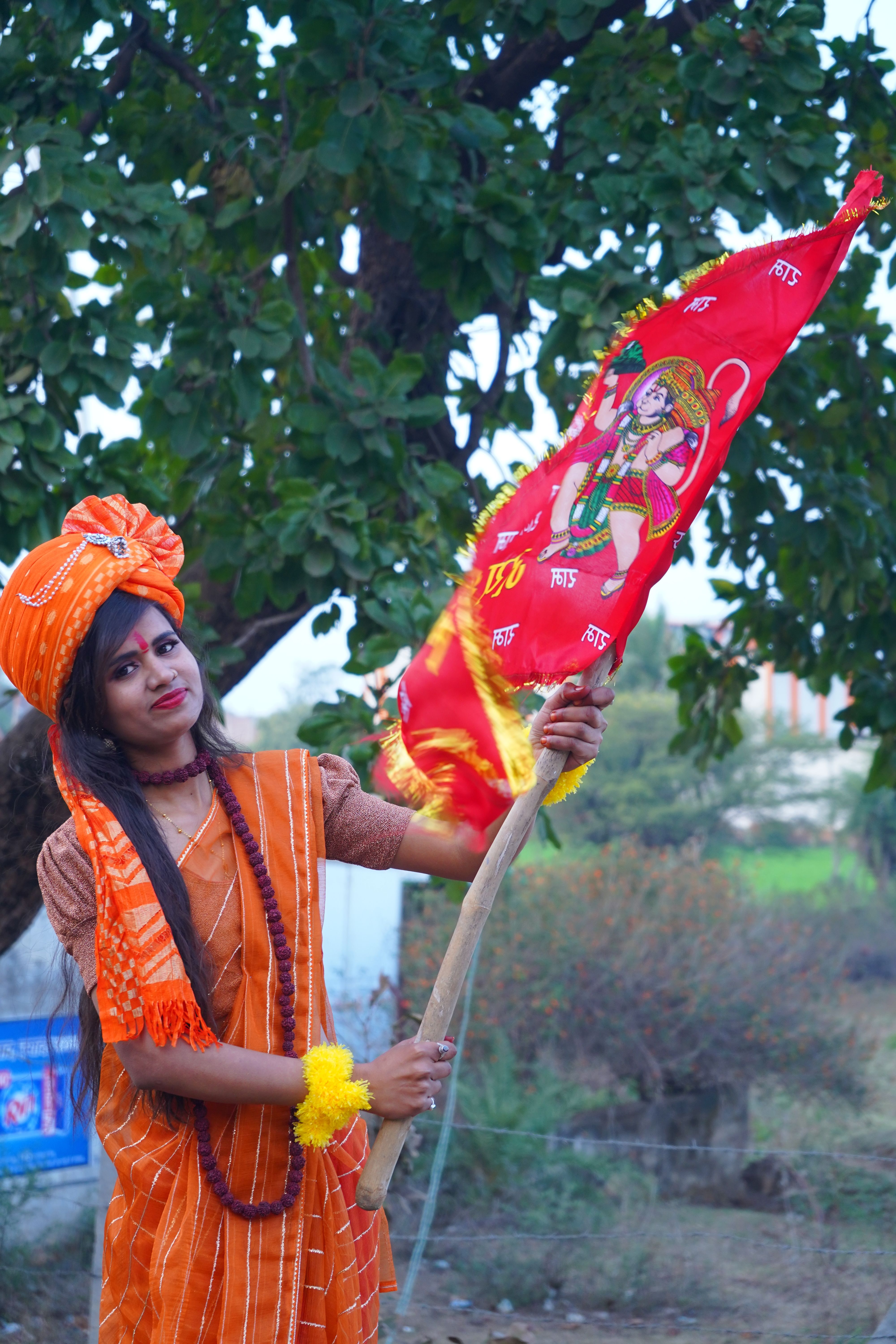 A South Asian woman in orange attire holds a colorful cultural flag outdoors, celebrating Indian heritage.