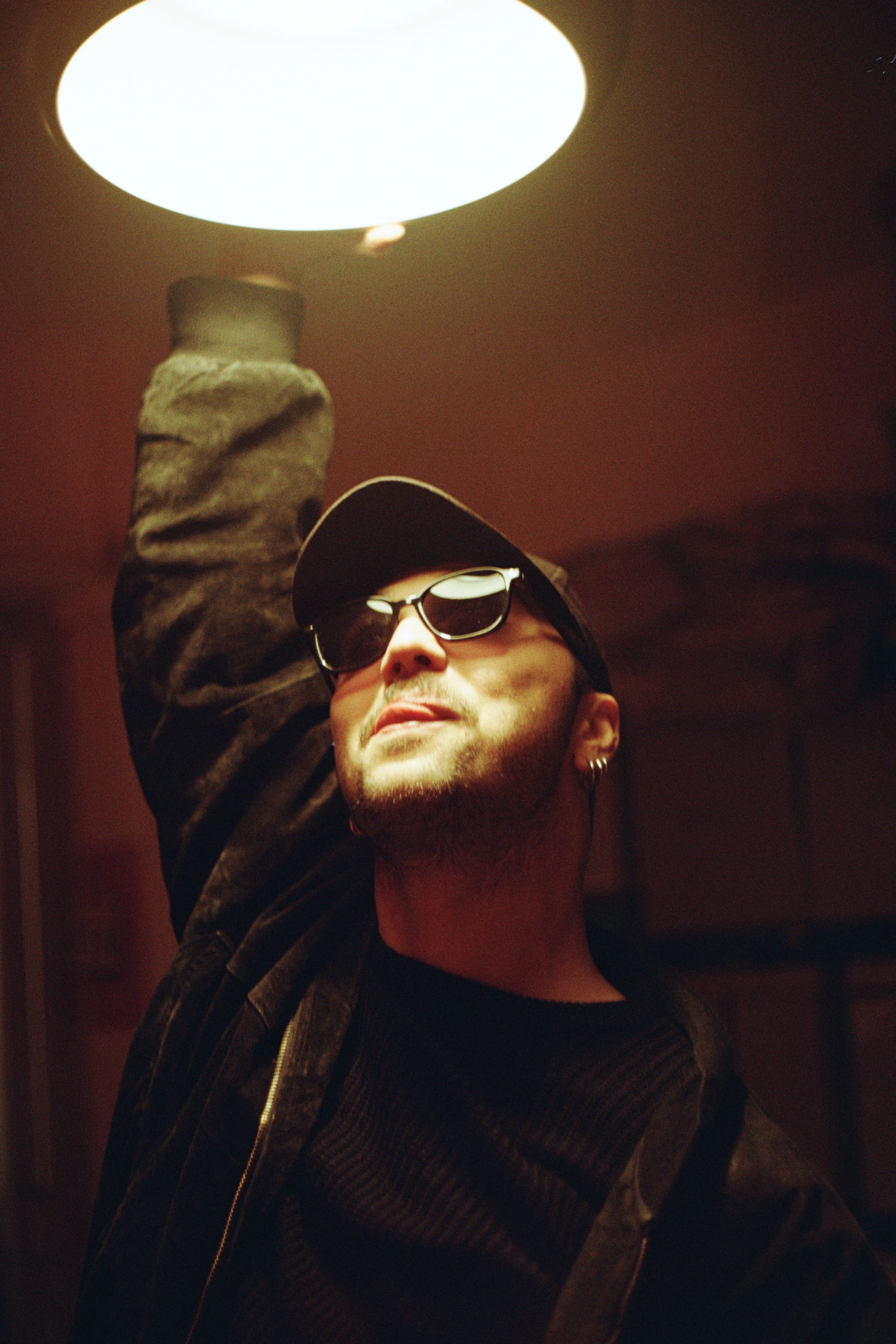 A man in sunglasses and cap reaches for a light, casting bold shadows indoors.