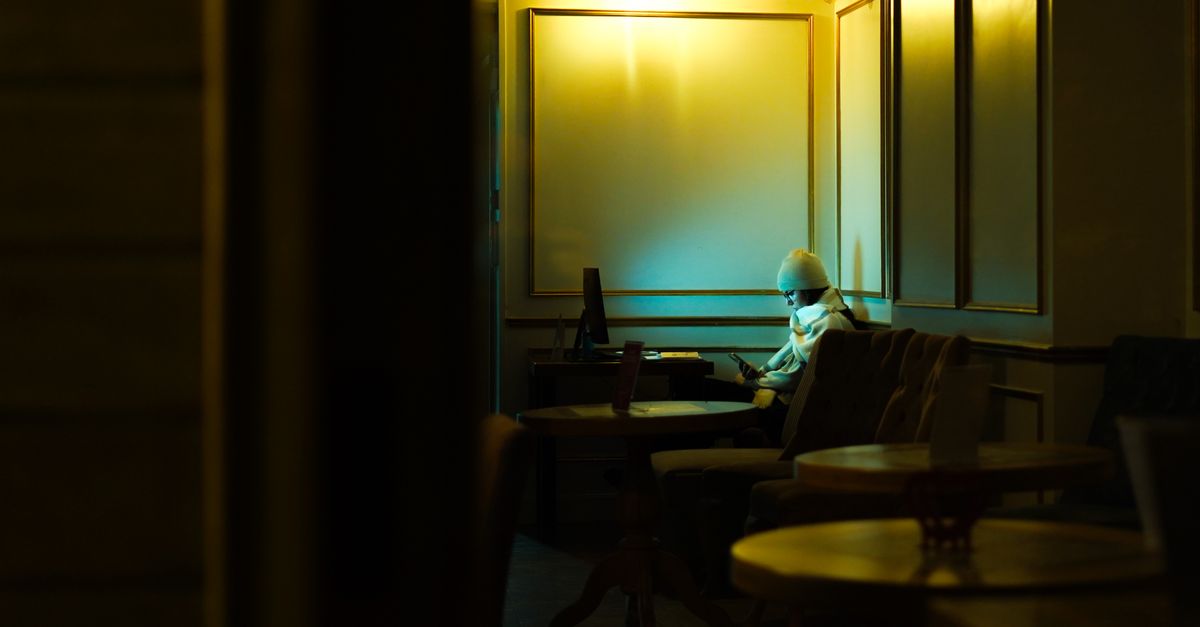 Uma pessoa solitária está sentada em um canto de café pouco iluminado, criando uma atmosfera sombria.
