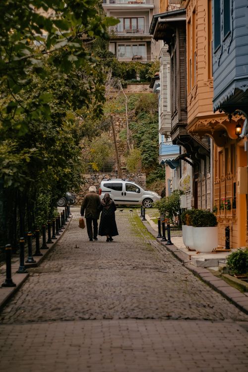 Free An elderly couple walking along a charming cobblestone street lined with colorful buildings. Stock Photo