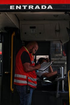 A transit worker in a red vest checks a clipboard while standing inside a city bus.
