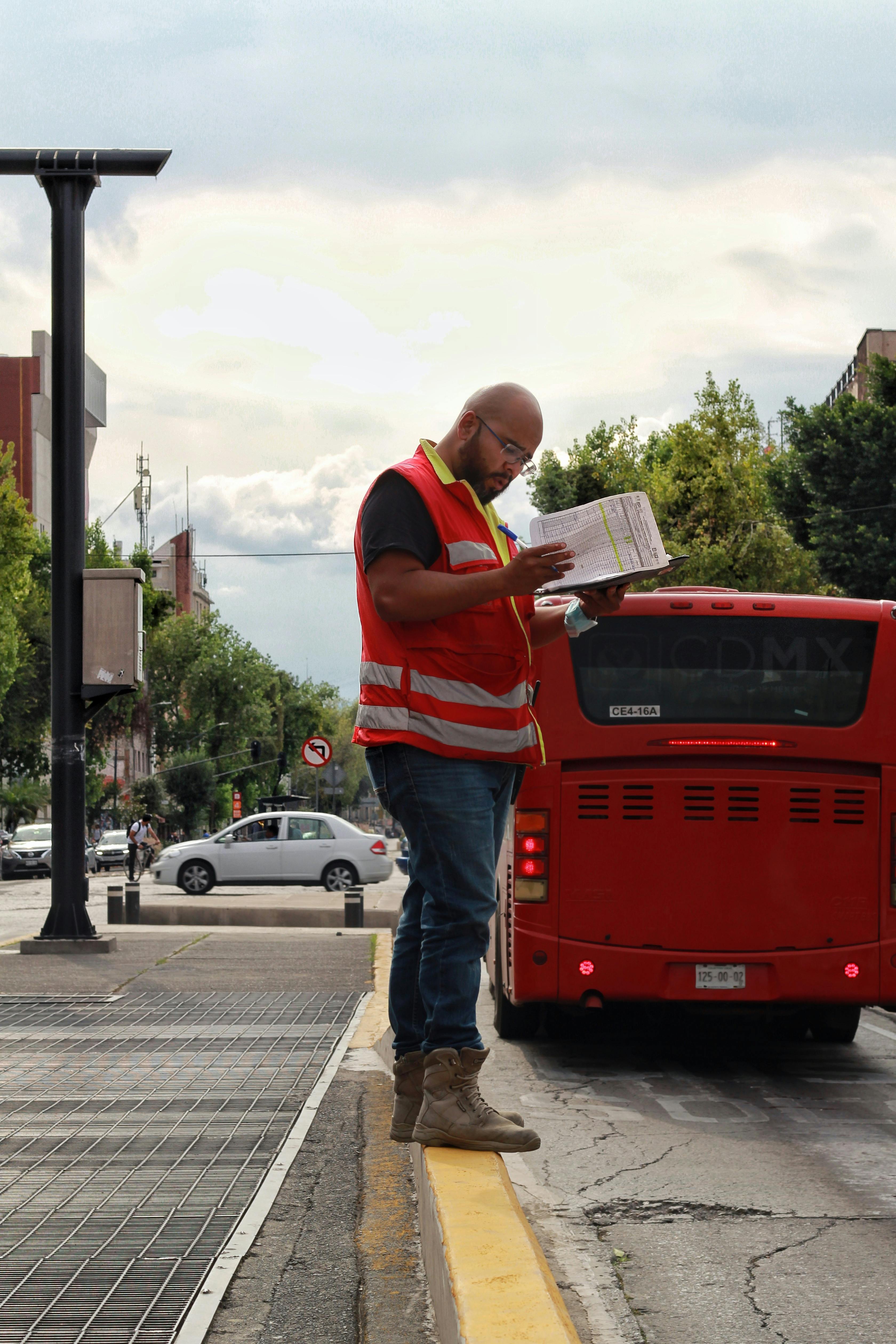 Man in reflective vest on city street, checking documents near a red bus.