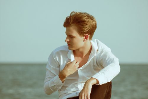 Free Young man in a white shirt sitting by the sea, lost in thought. Stock Photo
