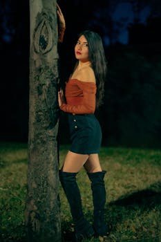 A stylish woman poses by a tree in a night city scene, wearing chic attire and boots, showcasing fashion in Mexico City.
