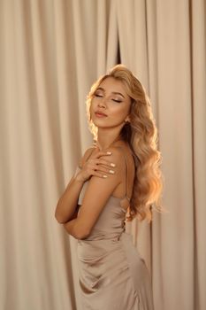 Glamorous woman with curled blonde hair in sleek satin dress posing gracefully with eyes closed.