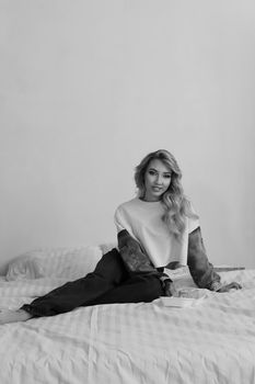 Elegant woman with long blonde hair sitting comfortably on bed in black and white fashion pose.