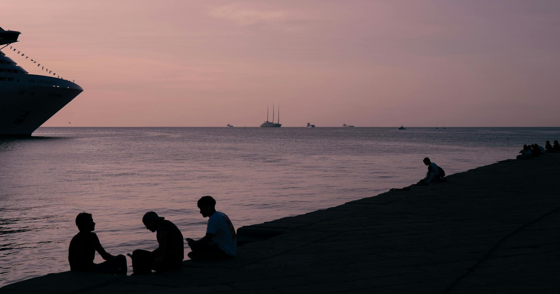 Silhouetted figures by the sea at sunset with ships in view at Trieste, Italy's waterfront promenade.