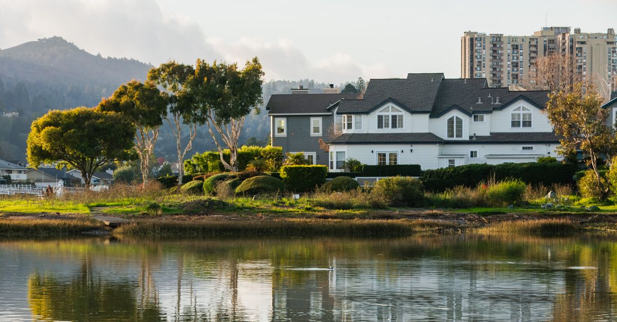 A peaceful suburban house by a serene lake with lush greenery and distant mountains.